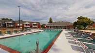 This two-story residential community is defined by its impressive outdoor amenity space, centered around a large swimming pool with striking brick-edged coping and clear turquoise water. The pool area is generously sized, accommodating a wide concrete sun deck lined with numerous lounge chairs, offering ample space for relaxation and recreation. A dedicated pool house and clubhouse building anchors one end of the complex, constructed in a classic brick style with a hipped roofline and a covered porch that provides shade and shelter. Adjacent to the pool, a raised terrace area with brick retaining walls offers additional seating, while a charming gazebo sits nearby, framed by lush greenery. The surrounding landscape features mature trees that provide natural shade and a pleasant visual backdrop. A sports court is also visible within the secured, fenced perimeter of the amenity area. The residential buildings themselves are two-story brick structures with pitched roofs, private balconies, and chimneys, reflecting a traditional architectural style. The overall ambiance is that of a well-appointed, community-focused property designed to support an active outdoor lifestyle.