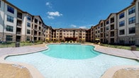 This residential unit is part of a well-appointed multi-story community characterized by a striking courtyard-style layout. The exterior showcases a combination of stone and wood-tone siding, with covered balconies lining each floor. At the heart of the community sits a generously sized resort-style pool with a freeform shape, surrounded by a spacious sundeck furnished with lounge chairs and yellow umbrella-shaded seating areas — ideal for outdoor relaxation.

The bedroom is thoughtfully designed with soft, neutral tones complemented by muted sage-green accent walls that lend a calm, cohesive atmosphere. Carpet flooring adds warmth underfoot, while a large window with horizontal blinds allows natural light to filter in, offering views of the surrounding community. White furniture pieces, including a dresser and nightstand, provide functional storage and a clean aesthetic contrast to the warmer wall tones.

A dedicated walk-in closet with built-in shelving and hanging rods offers organized storage space. An adjacent doorway leads directly into the en-suite bathroom, which features dark cabinetry and tile flooring. The overall layout is efficient and well-connected, creating a private and comfortable living environment within a vibrant residential community.