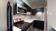 Located at 1075 Weybridge Rd in Columbus, Ohio, this single-floor residence offers a thoughtfully designed living space with modern finishes throughout. The kitchen serves as a standout feature, showcasing rich dark cabinetry with sleek hardware that creates a striking contrast against the crisp white countertops. A contemporary mosaic tile backsplash in neutral tones of gray and white adds texture and visual interest to the space. Stainless steel appliances, including a gas range and an over-the-range microwave, provide both functionality and a polished aesthetic. The double-basin stainless steel sink is positioned beneath under-cabinet lighting, enhancing both practicality and ambiance. Wood-look flooring runs through the kitchen, adding warmth and continuity to the design. The overall aesthetic of the home balances modern sophistication with everyday functionality, making it an ideal space for those who appreciate clean lines, quality finishes, and a well-organized layout. The single-floor configuration ensures easy accessibility and a seamless flow throughout the home.