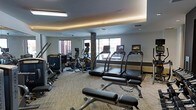 This fitness center is a thoughtfully designed, single-floor exercise facility that provides a comprehensive workout environment. The space features a tray ceiling with integrated ambient lighting that casts a warm, even glow throughout the room, complementing the recessed overhead lighting for a bright and energizing atmosphere. Large windows along the exterior wall allow natural light to fill the space while offering views of the surrounding area, creating an open and inviting feel.

The cardio section is prominently equipped with multiple modern treadmills, each outfitted with individual touchscreen display consoles, as well as elliptical cross-trainers to accommodate a variety of aerobic workouts. A dedicated strength and resistance training area occupies one side of the room, featuring cable machines, a weight bench with an adjustable rack, and additional strength equipment to support a full-body training regimen.

Decorative stone-clad columns add an architectural accent that lends texture and visual interest to the space. The carpeted flooring provides a comfortable, cushioned surface underfoot and helps define the exercise zones within the room. Overall, the fitness center is a well-appointed, functional space designed to support a wide range of fitness activities in a clean, modern setting.
