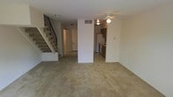 This two-story residence is centered around an open main living area that provides a functional and versatile layout. The living space is finished with wall-to-wall carpeting, contributing to a warm and comfortable atmosphere. A ceiling fan with a built-in light fixture is positioned overhead, providing both airflow and ambient lighting throughout the space.

A staircase with a dark metal railing runs along one side of the room, providing access to the upper floor. The staircase is carpeted to match the main level, creating a cohesive look throughout.

Toward the rear of the main level, the kitchen is partially separated by a structural dividing wall, creating a semi-open layout. The kitchen features tile-style flooring, dark wood cabinetry, and includes a refrigerator and stove. A secondary ceiling fan is installed in this area, ensuring good air circulation.

The front entry door is visible from the main living area, offering a straightforward and practical layout. The neutral wall tones and light-colored finishes throughout the space create a clean, move-in ready environment that allows for easy personalization.