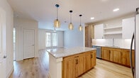 This thoughtfully designed 1-bedroom, 1-bathroom residence offers a seamless blend of modern aesthetics and functional living. The open-concept kitchen and living area serves as the heart of the home, anchored by a large center island with a granite countertop and striking amber glass pendant lights overhead. The kitchen is outfitted with warm wood and white cabinetry, stainless steel appliances, a tile backsplash, and ample storage including a tall pantry cabinet. Light wood-look flooring runs throughout the main living areas, lending a bright and cohesive feel. The living room receives natural light through a set of glass French doors that open to an outdoor space, complemented by a ceiling fan above. The bedroom is a comfortable retreat featuring plush carpet, a ceiling fan, and a window with outdoor views. A dedicated closet provides organized storage. The full bathroom is well-appointed with a soaking tub and tiled shower surround, a white vanity with built-in shelving, and wood-look flooring that ties back to the main living areas. The walk-in closet, accessible directly from the bathroom, adds everyday convenience. Clean white walls and recessed lighting throughout create a fresh, contemporary ambiance.