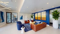 Located at 3300 Boutwell Rd in Lake Worth Beach, Florida, this single-floor space presents a thoughtfully designed lobby and living lounge area that blends contemporary architecture with vibrant interior design. The layout flows seamlessly from an entry foyer—featuring glass-paneled double doors, polished light tile flooring, and a striking large-scale abstract artwork in oceanic blues—into a welcoming reception area anchored by a sleek dark counter with pendant lighting overhead.

The central living area commands attention with a bold cobalt blue feature wall displaying a triptych of luminous, horizon-inspired paintings. Rich cognac leather sofas are paired with deep navy blue accent chairs and arranged around a circular wood coffee table atop a plush blue area rug, creating a cohesive and inviting color palette. Track and linear LED ceiling fixtures provide layered lighting throughout, complemented by a coffered ceiling detail that adds architectural depth.

Tall fiddle-leaf fig trees in white cylindrical planters introduce natural elements and vertical interest to the space. Glass-walled rooms along the perimeter allow visual connectivity while maintaining defined zones. The overall ambiance is polished and professional, with a coastal-modern aesthetic that reflects the South Florida setting.