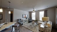 This single-floor residence offers a thoughtfully designed open-concept layout that seamlessly connects the living and dining areas. The living room is a standout feature, bathed in natural light streaming through multiple large windows dressed with elegant draping curtains and adjustable blinds. Smooth, light-toned walls and durable wood-look flooring create a clean, contemporary aesthetic throughout the space.

A ceiling fan with a stylish fixture anchors the living area, ensuring comfort year-round. The room accommodates a wall-mounted flat-screen television above a white media console with glass-front cabinetry, providing both functional storage and visual appeal. Comfortable seating — including a plush sofa, upholstered accent chairs, and a soft, textured area rug — establishes a cozy yet refined atmosphere. A glass-top coffee table with metallic legs adds a modern, airy touch to the seating arrangement.

Adjacent to the living area, a bar-height countertop with seating hints at an open kitchen connection, ideal for casual dining and entertaining. The neutral color palette and carefully selected furnishings lend the space a balanced, inviting ambiance that is both functional and stylish, making it well-suited for comfortable everyday living.