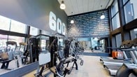 This fitness center is a thoughtfully designed workout facility that combines functionality with motivational aesthetics. The space is anchored by a bold, dark feature wall emblazoned with large-scale motivational phrases — including "Set Goals," "Push Yourself," and "No Excuses" — alongside a prominent "600" graphic, creating an energizing atmosphere from the moment you enter. Floor-to-ceiling mirrors line one side of the room, visually expanding the space and allowing members to monitor their form during workouts. The facility is outfitted with a comprehensive selection of strength-training machines, including multi-function cable and resistance equipment, providing a well-rounded workout experience. Cardio equipment, including treadmills, is visible toward the rear of the space. Modern pendant lighting hangs from the ceiling, providing focused illumination throughout the gym floor. The light-toned flooring contrasts sharply with the dark walls, contributing to a sleek and contemporary visual balance. Overall, the space delivers a professional, high-energy environment designed to keep members motivated and focused on their fitness goals.