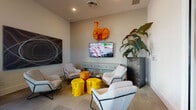 Located at 1450 Neo Square Blvd in Kissimmee, Florida, this single-floor community amenity space is thoughtfully designed to serve as a social and recreational hub. The interior is defined by a contemporary aesthetic, with neutral wall tones and light flooring that create a clean, modern backdrop throughout.

The lounge area is a focal point of the space, furnished with sleek, upholstered accent chairs arranged around vibrant yellow stool-style tables. A wall-mounted flat-screen television, a bold orange elephant wall sculpture, and a large abstract painting add artistic personality to the room. A towering indoor plant in a textured planter introduces a natural element, while a console table beneath the TV grounds the arrangement.

Adjacent to the lounge, a billiards area offers a full-size pool table set beneath industrial-style pendant lighting, accompanied by additional lounge seating and coordinating yellow accent stools. Abstract wall art continues the design language established throughout the space.

The dining nooks are bathed in natural light, courtesy of expansive floor-to-ceiling windows that frame views of the outdoor pool area and surrounding landscaping. Warm wood round tables paired with matching chairs create an inviting atmosphere ideal for casual gatherings or meetings. Curated artwork punctuates the walls, adding visual interest to each corner of this well-appointed community space.
