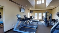 This fitness center is designed to offer a comprehensive workout environment within a single-floor layout. The space is anchored by a row of Matrix brand treadmills and cardio machines, positioned to take advantage of the natural light streaming in through several large windows along the walls. The elevated ceiling with a raised tray detail adds a sense of openness and volume to the room, preventing any feeling of confinement despite the focused purpose of the space. Multiple wall-mounted flat-screen televisions are strategically placed for visibility from the cardio equipment, enhancing the workout experience. Strength training equipment is positioned toward the periphery of the room, allowing for a logical and spacious flow between cardio and resistance training areas. The flooring is a durable, textured carpet suited for a fitness environment, and recessed lighting complements the natural light to keep the space well-illuminated at all times. The warm neutral tones of the walls, paired with the dark crown molding, lend the fitness center a polished and welcoming ambiance that balances functionality with an inviting aesthetic.