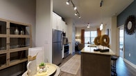 This well-appointed single-floor residence showcases a thoughtfully designed open-concept layout that seamlessly connects the kitchen and living areas. The kitchen is a standout feature, equipped with stainless steel appliances including a refrigerator, microwave, and range, all complemented by crisp white cabinetry that extends to the ceiling for ample storage. Rich granite countertops line the kitchen island, which also functions as a casual dining and prep space. Track lighting runs the length of the kitchen ceiling, providing bright, directed illumination throughout the cooking area. The hardwood-style flooring extends from the kitchen into the living space, creating a cohesive and clean aesthetic. The living area benefits from generous natural light streaming through large windows dressed with warm-toned curtains, lending the space an inviting ambiance. A ceiling fan adds both style and function overhead. The neutral color palette of warm grays and whites throughout creates a calm, modern atmosphere that feels both spacious and comfortable. Decorative pendant lighting over the island adds an elegant finishing touch, tying together the contemporary design of this well-designed residence.