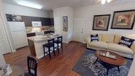 This single-floor, one-bedroom, one-bathroom unit offers a thoughtfully organized layout that maximizes its compact footprint. The open-concept living and kitchen area serves as the heart of the unit, anchored by rich hardwood-style flooring that runs throughout. Crown molding adds a refined architectural detail to the main living space, while a ceiling fan provides both function and style.

The kitchen is equipped with dark cabinetry, white appliances including an electric range and over-the-range microwave, and light-toned countertops. A peninsula breakfast bar with seating for two bridges the kitchen and living area, offering a practical dining and social space. The living room benefits from a glass patio door that invites natural light and provides outdoor access.

The bedroom is a comfortable, well-proportioned retreat featuring a ceiling fan, a window with curtains allowing natural light, and hardwood-style flooring consistent with the rest of the unit.

The bathroom features a full soaking tub with a shower curtain, a vanity with dark cabinetry, and the same warm flooring found throughout. Additional storage is accessible via a closet adjacent to the bathroom, contributing to the unit's overall practicality.