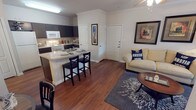 This single-floor, one-bedroom, one-bathroom unit offers a thoughtfully organized layout that maximizes its compact footprint. The open-concept living and kitchen area serves as the heart of the unit, anchored by rich hardwood-style flooring that runs throughout. Crown molding adds a refined architectural detail to the main living space, while a ceiling fan provides both function and style.

The kitchen is equipped with dark cabinetry, white appliances including an electric range and over-the-range microwave, and light-toned countertops. A peninsula breakfast bar with seating for two bridges the kitchen and living area, offering a practical dining and social space. The living room benefits from a glass patio door that invites natural light and provides outdoor access.

The bedroom is a comfortable, well-proportioned retreat featuring a ceiling fan, a window with curtains allowing natural light, and hardwood-style flooring consistent with the rest of the unit.

The bathroom features a full soaking tub with a shower curtain, a vanity with dark cabinetry, and the same warm flooring found throughout. Additional storage is accessible via a closet adjacent to the bathroom, contributing to the unit's overall practicality.