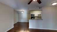 This single-floor space is designed with an open-concept layout that seamlessly connects the living area and kitchen, creating a cohesive and functional environment. Rich, dark hardwood flooring runs throughout, providing a warm and grounded aesthetic that complements the neutral, light-toned walls.

The kitchen is positioned within a raised nook defined by clean white cabinetry that extends to the ceiling, offering ample storage. A granite-topped breakfast bar peninsula serves as both a workspace and a casual dining surface, bridging the kitchen and living areas. Stainless steel appliances, including a refrigerator, microwave, and a black range, lend a modern, polished finish to the culinary space.

The living area is open and airy, illuminated by a combination of a ceiling fan with an integrated light fixture and a decorative pendant light. These overhead lighting elements contribute to the overall brightness and comfort of the space. A closet door is visible along one wall, adding practical storage within the living zone.

The layout, finishes, and fixtures work together to create a clean, contemporary atmosphere that is both welcoming and well-appointed. The thoughtful design balances style and practicality, making the space highly functional for everyday living.