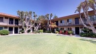 This inviting courtyard-style property is centered around a generous, well-manicured lawn that serves as the heart of the community. The two-story building wraps around the outdoor space in a U-shaped configuration, with each level featuring private balconies that overlook the verdant grounds below. The warm, neutral stucco exterior is accented by a classic Spanish-inspired red tile roofline, lending the property a timeless Southern California character.

The landscaping is a standout feature, showcasing a rich variety of tropical and Mediterranean plantings. Towering banana trees, mature eucalyptus with striking sculptural trunks, fan palms, and vibrant flowering shrubs create a layered, resort-like atmosphere throughout the courtyard. Concrete walkways wind through the lush greenery, connecting different sections of the building while offering a pleasant stroll through the botanical surroundings.

Seating areas with red Adirondack chairs are tucked among the plantings, inviting residents to relax and enjoy the outdoor ambiance. The ground-floor units open directly to the courtyard via sliding glass doors, while upper-level units benefit from elevated balcony views. Under sunny blue skies, the entire property radiates a bright, warm, and welcoming character that reflects its Southern California setting.
