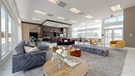 This expansive single-floor space is designed with a modern, upscale aesthetic that seamlessly integrates multiple functional areas within an open-plan layout. Polished marble-look porcelain tile flows throughout, establishing a refined foundation that unifies the entire space. Soaring ceilings fitted with recessed panel lighting amplify the sense of volume, while floor-to-ceiling windows and clerestory glazing along multiple walls flood the interior with abundant natural light and provide sweeping outdoor views of the surrounding greenery and pool areas.

The main lounge area features an array of curated contemporary furnishings, including deep velvet sofas, tufted ottomans, and accent chairs in warm gold tones, all anchored by plush area rugs. A feature wall with textured paneling and a mounted television provides a focused media zone. Adjacent to the lounge, a generous dining area accommodates a large rectangular table with seating for ten or more, positioned directly in front of expansive windows framed by sheer drapery.

The kitchen zone is anchored by a dark-toned cabinetry system with open shelving, a long white stone bar counter with bar seating, and gold pendant lighting overhead. A casual banquette seating area nearby overlooks the outdoor pool, providing a relaxed alternative gathering space. The overall ambiance is polished, bright, and welcoming.