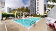 Located at 1425 Bedford St in Stamford, Connecticut, this outdoor amenity space centers around a generously sized swimming pool with crystal-clear blue water. The pool area is thoughtfully designed for both relaxation and social gathering, offering a well-maintained concrete deck that surrounds the pool on all sides. A striking white pergola structure runs along one side of the pool, providing partial shade over a row of lounge chairs — ideal for sunbathing while enjoying filtered natural light. Additional seating options include Adirondack-style chairs and an umbrella-shaded dining table set, accented by vibrant potted flowers that add a welcoming touch of color. A sleek, modern glass-fronted clubhouse or amenity building is positioned at one end of the pool, featuring floor-to-ceiling windows that seamlessly connect the indoor and outdoor spaces. Neatly trimmed arborvitae hedges and mature trees line the perimeter, providing privacy and a lush green backdrop against the open sky. The space is enclosed by a secure fence and is set adjacent to a multi-story residential tower, situating this amenity area within an established urban residential community in Stamford, CT.
