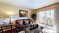 This single-floor residence is designed with a cohesive, warm aesthetic carried throughout its living spaces. The living room serves as a central gathering area, featuring rich hardwood-style flooring, a chair rail accent along the walls, and sheer patterned drapes framing a large sliding glass door. The door opens directly onto a private balcony, allowing natural light to fill the room and providing an outdoor seating area. A decorative area rug and wood-framed coffee table anchor the space.

The dining area is positioned adjacent to the living room and continues the hardwood-style flooring. It includes a pass-through window connecting to the kitchen, a framed wall mirror, and a closet for additional storage. The kitchen is outfitted with white cabinetry, dark granite-style countertops, a tile backsplash, and black appliances, including a range with overhead ventilation.

The bedroom offers plush carpeting, a window with blinds and curtains, and sufficient space for a full bedroom suite. Walls throughout the residence are painted in a soft warm beige, contributing to the overall inviting and comfortable atmosphere of the home.