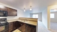 This single-floor space offers a well-designed open-concept layout that seamlessly connects the kitchen and living area. The kitchen is outfitted with rich, dark cabinetry that provides ample storage, complemented by laminate countertops and a double-basin stainless steel sink. A built-in dishwasher adds everyday convenience, while the countertop peninsula creates a natural visual boundary between the kitchen and living space. Overhead, two pendant light fixtures with classic globe shades provide focused task lighting and contribute to the space's clean aesthetic. The flooring transitions from hard surface in the kitchen to plush carpet in the living area, offering a comfortable and practical distinction between the two zones. The living area benefits from a large sliding glass door that fills the room with natural light and provides an outdoor connection. Neutral wall tones and a light-colored ceiling throughout lend a bright, airy quality to the entire space. The overall design is functional and approachable, making it well-suited for everyday living.