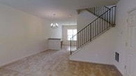 This well-appointed three-story residence offers a thoughtfully designed layout spread across multiple levels, providing ample space for comfortable living. The main living area greets you with neutral-toned carpeting that lends a warm, inviting atmosphere throughout the open floor plan. A striking chandelier anchors the living space, casting a soft glow over the room, while a second pendant light illuminates the adjoining area near a bright window that brings in natural light. A raised breakfast bar serves as a natural dividing point between the living space and the kitchen area, offering a functional and stylish transition between the two zones. The staircase, featuring sleek black iron balusters and a clean white stringer, adds a contemporary architectural touch and connects the floors with visual elegance. The neutral color palette of white walls and light carpeting throughout creates a bright, airy ambiance that offers a versatile canvas for a variety of interior design styles. With three full floors of living space, this residence provides generous room for both private retreats and shared living, making it a practical and refined home for everyday life.