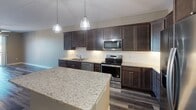 This well-appointed single-floor residence in Wausau, Wisconsin, offers a thoughtfully designed layout with contemporary finishes and quality materials throughout. The kitchen serves as a standout centerpiece, showcasing rich dark-stained cabinetry, light granite countertops, a large island, and a full suite of stainless steel appliances including a range, microwave, dishwasher, and refrigerator with ice maker. Industrial-style pendant lighting adds a modern touch above the island, while wood-look plank flooring grounds the space with warmth and durability. The open living area adjacent to the kitchen features the same plank flooring, creating a seamless flow and a spacious, airy feel. The bedroom is generously sized, appointed with plush carpet, a ceiling fan with light fixture, and a large window that fills the room with natural light and offers open outdoor views. The bathroom continues the cohesive design aesthetic with granite-topped dark cabinetry and hardwood-style flooring that transitions into carpeted bedroom and hallway areas. The unit also includes a dedicated laundry area, adding everyday convenience. Neutral wall tones throughout complement the modern finishes, creating a clean, move-in-ready environment.