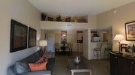This single-floor residence showcases an open-concept design that seamlessly connects the main living spaces. The living room is a standout feature, boasting impressively high ceilings with an elevated display ledge that adds architectural character and vertical depth to the space. Warm, neutral tones throughout create a welcoming and cohesive ambiance.

The living area is generously proportioned, providing ample room for comfortable seating arrangements centered around a coffee table. It flows naturally into a dedicated dining area, which features a round dining table with seating for multiple guests, making it ideal for both casual meals and entertaining.

The kitchen is efficiently designed with light-toned cabinetry, modern appliances including a refrigerator, and a breakfast bar with tall bar seating — offering a convenient spot for quick meals or conversation. The kitchen's open connection to the main living and dining areas enhances the sense of togetherness throughout the home.

Carpeted flooring extends through the main living spaces, contributing to the warm and inviting feel of the interior. The layout is practical and well-proportioned, with clearly defined zones that still maintain an airy, open atmosphere throughout the single-level floor plan.