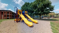 Located at 2255 Country Corners Dr in Columbus, Ohio, this single-level outdoor recreational space centers around a bold, colorful playground structure designed to engage and entertain. The play equipment features a striking combination of red, blue, and yellow components, highlighted by two prominent yellow slides that extend outward from a central blue tube structure. The framework is supported by red and blue posts, and the play unit incorporates additional interactive elements such as a spinning wheel panel, offering a variety of activities for children. The ground beneath the structure is covered in a layer of protective wood chip surfacing, providing both safety and a natural aesthetic underfoot. Surrounding the playground, a canopy of large, mature trees offers generous shade and a pleasant natural setting. A picnic table area is visible nearby, providing a space for gathering and relaxation alongside the play zone. The open layout of the grounds allows for ease of movement and supervision throughout the space. The overall environment is bright, welcoming, and well-suited for active outdoor use, combining durable equipment with a comfortable, tree-lined setting.