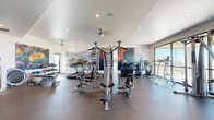 Located at 3510 N Craycroft Rd in Tucson, Arizona, this fitness center offers a thoughtfully designed, single-floor workout space with a generous open layout. The interior is finished with durable dark rubber flooring throughout, recessed ceiling lighting, and ceiling fans that maintain a comfortable environment during workouts.

The facility is comprehensively equipped for both strength and cardiovascular training. A multi-station cable machine serves as the centerpiece, complemented by a dumbbell rack, benches, and an assortment of resistance equipment. Cardio offerings include multiple treadmills, stationary bikes, elliptical trainers, and a stair climber. A dedicated stretch and functional training area features stability balls, foam rollers, and a water station.

One of the most distinctive design elements is the large-scale desert landscape wall murals, depicting iconic Sonoran Desert scenes — including saguaro cacti, mountain vistas, and vivid Arizona sunsets — creating an immersive, regionally inspired atmosphere. Floor-to-ceiling windows and sliding glass doors along one wall flood the space with natural light and provide sweeping views of the surrounding Tucson landscape. The combination of vibrant artwork, natural light, and high-quality equipment makes this fitness center both functional and visually engaging.
