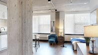 Located at 708 22nd St in Greeley, Colorado, this studio unit showcases an industrial-modern aesthetic defined by exposed concrete ceilings, structural concrete columns, and warm hardwood flooring throughout. The open-plan layout makes efficient use of the space, with a decorative lattice-panel room divider helping to delineate the sleeping area from the main living zone without sacrificing the airy, loft-like feel. Track lighting runs along the ceiling, complementing the natural light that pours in through large windows fitted with horizontal blinds. The kitchen is well-appointed with white shaker cabinetry, stone-look countertops, and a full suite of stainless steel appliances including a refrigerator, electric range, microwave, and dishwasher. A bistro-style table and stools provide a casual dining option within the kitchen area. The bathroom features a full soaking tub and shower combination with clean white subway tile surround, accessed through a bold black interior door that adds a sharp contrast to the lighter finishes. Two closets offer practical storage solutions. The unit's industrial character, combined with contemporary finishes, creates a cohesive and functional living environment with strong visual appeal.