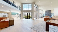 Located on the 3rd and 4th floors at 100 Avenue G in Bayonne, NJ, this impressive two-story amenity space offers a generous and versatile layout across two levels. The main floor commands attention with soaring double-height ceilings and floor-to-ceiling black-framed windows that flood the interior with abundant natural light and provide open views of the surrounding neighborhood. Light-toned hardwood floors run throughout, complementing the bright, airy aesthetic.

The kitchen is outfitted with sleek light wood cabinetry accented by brass hardware, a herringbone-patterned white tile backsplash, a large island with bar seating, and open shelving — creating a functional and stylish gathering hub. Adjacent to the kitchen, the expansive living area features multiple lounge configurations, including a media lounge with a large screen, plush seating, and warm wood built-ins.

A dramatic spiral staircase leads to the upper level, where a carpeted game room houses a shuffleboard table, a card table, and high-top bar seating. A separate area on this level also includes a pool table set against wainscoted walls. A hallway connects the various zones seamlessly. The teal accent walls and curated artwork throughout add a modern, residential warmth to this thoughtfully designed communal space.