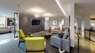 This thoughtfully designed residential amenity floor offers a seamless blend of contemporary architecture and curated interior design. The grand lobby entrance makes an immediate impression with soaring double-height ceilings, expansive floor-to-ceiling windows, and large-format tile flooring that draws in an abundance of natural light. Vibrant chartreuse accent chairs arranged around a sculptural round coffee table create an inviting seating area with views to the outdoors.

Adjacent to the entrance, a well-appointed mailroom and package area features a sleek marble-topped counter with under-cabinet storage and recessed cove lighting overhead. Nearby, a designated E-Lounge offers a cozy booth with white upholstered banquette seating, a warm wood-toned table, and eye-catching geometric patterned wallcovering in soft mauve tones, anchored by a statement pendant light.

The main lounge area is a standout social hub, combining a richly textured dimensional accent wall with a wall-mounted flat-screen TV, distressed wood-look wall panels, and layered ambient lighting from globe pendants and cove fixtures. Plush sectional sofas in neutral grey tones are complemented by bold yellow accent chairs, curated artwork, and a collection of framed geometric prints, creating a dynamic yet comfortable gathering space for residents.