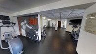 The Artisan Fit Club is a single-level fitness center designed to provide a functional and motivating workout environment. The space is immediately distinguished by a striking black-and-white Muhammad Ali mural emblazoned with the iconic phrase "Sting Like A Bee," serving as a bold focal point that energizes the atmosphere. The room features a drop-panel ceiling with recessed and fluorescent lighting, ensuring consistent, even illumination throughout.

The fitness floor is finished with dark rubber flooring, a practical and durable surface well-suited for high-traffic exercise use. Cardio equipment, including upright stationary bikes and an elliptical machine, is positioned along one side of the room, accompanied by a wall-mounted flat-screen television for entertainment during workouts. On the opposite side, a selection of strength training machines and free-weight equipment provides options for resistance training. A wooden storage unit is visible toward the rear of the space, adding a warmer visual element. Posted gym rules are displayed near the entrance, indicating structured, community-oriented use of the facility.