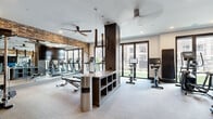 This modern fitness center offers a thoughtfully designed workout space that blends industrial and contemporary aesthetics. Exposed brick walls provide a striking backdrop throughout, complemented by light-toned carpet flooring and crisp white ceilings fitted with recessed lighting and sleek ceiling fans for air circulation. A dark wood central divider column adds architectural interest while subtly organizing the space into distinct workout zones. The cardio area features a row of treadmills and elliptical machines positioned along a large mirrored wall, which amplifies the sense of openness and allows users to monitor their form. A rowing machine and stationary bikes with integrated display screens round out the cardio offerings. The strength training zone is equipped with a Matrix cable machine, free-weight benches, and a well-stocked dumbbell rack. Natural light floods the space through expansive floor-to-ceiling windows and glass sliding doors, offering views of a landscaped outdoor area. The entrance is framed by warm wood-paneled walls and glass double doors, creating an inviting and polished entry point. Pendant lighting and wall-mounted flat-screen TVs add modern convenience, making this fitness center a fully equipped and visually engaging environment for a comprehensive workout experience.
