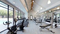 This fitness center offers a thoughtfully designed workout environment that combines architectural character with practical functionality. The space is defined by its dramatic vaulted ceiling, which features exposed wooden beams and industrial-style ductwork, lending a modern yet organic aesthetic. Pendant lighting hangs at varying heights, complementing the recessed ceiling lights to ensure the space is well-illuminated throughout.

A row of large, dark-framed windows lines one wall, flooding the interior with natural light and providing views of the surrounding greenery, creating an inviting atmosphere for exercise. The light-toned flooring with subtle accent striping adds a clean, contemporary look underfoot.

The cardio section is well-stocked with a row of treadmills, ellipticals, and stationary bikes from the TRUE Fitness brand, all positioned to take advantage of the natural light. On the opposite side, a dedicated strength training area includes cable machines, a multi-station weight system, and a free weight bench. A mirrored wall expands the sense of space and allows for proper form checks during workouts. Overall, the fitness center balances form and function seamlessly.
