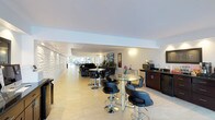 This contemporary single-floor lounge space is designed with a clean, modern aesthetic that prioritizes both comfort and functionality. The open-plan layout stretches generously, creating a spacious and airy environment filled with natural light streaming in from large windows at the far end.

The interior is anchored by a striking textured 3D accent wall along one side, adding depth and visual interest to the otherwise crisp white walls and ceilings. Polished light-toned floor tiles run throughout, contributing to the bright and welcoming ambiance.

Seating is arranged throughout the space with sleek black bar-height tables paired with contemporary swivel chairs, offering a modern and social atmosphere ideal for gatherings or casual meetings. Additional lounge seating is visible toward the rear, providing a more relaxed setting.

Along one wall, a dedicated kitchenette area is equipped with a countertop, sink, microwave, and a mini-refrigerator, making the space self-sufficient and convenient for guests or staff. Wall-mounted sconces and a flat-screen display add to the polished, hospitality-oriented feel of the environment.

Decorative touches, including potted plants and framed artwork, are thoughtfully placed throughout, lending warmth and personality to the otherwise minimalist design. Overall, the space presents a refined, professional atmosphere well-suited for a hotel lobby, corporate lounge, or upscale common area.