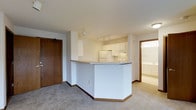 This single-floor residence offers a clean, functional layout designed to maximize everyday living. The open-concept kitchen is a central highlight, featuring white cabinetry, white appliances, and a peninsula countertop that creates a natural divide between the kitchen and the adjoining living area. Track lighting overhead illuminates the workspace efficiently, while the light-toned countertops and crisp white finishes contribute to a bright, airy atmosphere throughout. The living area is finished with plush carpeting in a neutral tone, providing a comfortable and cohesive feel. A solid wood entry door with warm stained trim welcomes residents into the space, which is accented throughout by coordinating wood baseboards and door frames. A glimpse into an adjacent room reveals additional white cabinetry and light tile flooring, suggesting a well-appointed bathroom or utility space nearby. The unit benefits from views of a peaceful, tree-lined neighborhood streetscape, where mature evergreen and deciduous trees frame well-maintained lawns and quiet residential streets. Overall, the space presents a tidy, move-in-ready environment with a practical design, neutral palette, and a calm suburban outlook.