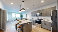 This thoughtfully designed one-bedroom, one-bathroom residence offers a smart and efficient layout across a single floor. The open-concept kitchen and living area serves as the heart of the home, featuring gray shaker-style cabinetry, white subway tile backsplash, stainless steel appliances, and a central island with seating — all illuminated by stylish pendant lighting and recessed fixtures. Light hardwood-style flooring runs continuously throughout, enhancing the sense of cohesion and warmth.

The living area flows naturally toward large windows and a glass door that invite abundant natural light and views of the surrounding exterior. A dedicated dining nook provides an ideal spot for casual meals or relaxed conversation.

The bedroom is a comfortable retreat with multiple windows offering outdoor views, a ceiling fan, and direct access to a spacious walk-in closet. The bathroom is finished with a gray vanity, white quartz countertop, an illuminated mirror, and a subway-tiled tub-and-shower combination with matte black fixtures. Additional conveniences include an in-unit laundry area and two closets for ample storage. The overall aesthetic is modern and clean, with neutral tones and contemporary details throughout.