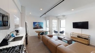 This contemporary single-floor unit showcases a thoughtfully designed open-concept layout that seamlessly connects the living, dining, and kitchen areas. The living space is anchored by a generous sectional sofa in warm cognac leather, a wood-and-glass coffee table, and a wall-mounted flat-screen TV set on a minimalist wood media console. Two accent chairs positioned near the large windows invite natural light to fill the room, while bold wall art adds a vibrant focal point to the dining area, which features a rustic wood dining table and eclectic seating. Exposed industrial ductwork and black track lighting run along the ceiling, blending urban character with modern sensibility. Wood-toned flooring throughout the main living area adds warmth and continuity. The kitchen is efficiently designed with stainless steel appliances, including a gas range and over-the-range microwave, complemented by two-tone cabinetry in white and natural wood, and a clean tile backsplash. The bedroom is a calm, understated retreat featuring plush carpet, a wood-framed bed, a built-in closet with sliding doors, and a simple bedside setup. The bathroom is crisp and minimal, outfitted with a deep soaking tub, shower, and brushed gold fixtures against bright white walls.
