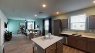 This thoughtfully designed single-floor residence offers a seamless open-concept layout that connects the kitchen, dining, and living areas into one cohesive and functional space. The kitchen is a standout feature, equipped with stainless steel appliances including a gas range, microwave, and refrigerator, complemented by grey cabinetry, light quartz countertops, and a central island that doubles as a casual dining and prep area. Wood-look flooring runs throughout the kitchen and entry spaces, adding warmth and durability.

The living room is finished in a soothing sage green palette with plush carpeting, large windows dressed with dark curtain panels that invite generous natural light, and a ceiling fan overhead. The space feels open and airy, with room for both a seating and dining arrangement.

The bedrooms are comfortably sized with soft carpet flooring and ceiling fans. The primary bedroom includes a walk-in closet and direct sightlines to the kitchen. A second bedroom features a bright window with exterior views. The bathroom is clean and functional, featuring a combination tub and shower, wood-look tile flooring, and a vanity with matching grey cabinetry and white countertop. Overall, the residence presents a cohesive, modern aesthetic throughout.