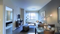This single-floor residence showcases a thoughtfully designed open-concept living area that blends contemporary style with functional comfort. Dark hardwood flooring runs throughout, providing a sleek foundation that contrasts elegantly with the light grey walls and white ceiling. A large window dressed in sheer white curtains allows abundant natural light to flood the room, creating an airy and inviting atmosphere.

The living area is anchored by a plush, neutral-toned sectional sofa adorned with decorative pillows in warm earth tones. A low-profile glass and metal coffee table sits atop a textured area rug, complemented by a woven cube ottoman that adds a tactile element to the space. A wall-mounted flat-screen television is paired with a dark wood media console providing organized storage below.

Decorative touches include a large abstract canvas artwork on the feature wall and a sleek floor lamp that adds ambient lighting. Potted plants near the window introduce a natural element, while a glass dining table visible to the side suggests a seamlessly integrated dining area. Overall, the space presents a modern, cohesive aesthetic.