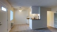 This single-story residence offers a practical and well-organized layout designed to maximize the use of its living space. The open-concept design connects the living area and kitchen, creating a cohesive and functional floor plan. The kitchen features white cabinetry, dark granite-style countertops, and a peninsula with a pass-through opening that looks out into the living area, making the space feel connected and communicative. Under-cabinet lighting illuminates the workspace, adding both functionality and warmth to the kitchen area. The living area is finished with plush carpeting, providing comfort underfoot, while the kitchen and adjoining transition areas feature tile flooring for easy maintenance. A ceiling fan with integrated lighting is centrally positioned in the living area, offering both air circulation and ambient illumination. The walls throughout are painted in a neutral, light tone, contributing to a clean and airy atmosphere. A textured ceiling adds a subtle design element consistent throughout the space. Natural light filters in through a window in the living area, brightening the interior and enhancing the overall openness of the single-floor layout.