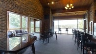 This single-floor space showcases a compelling mix of rustic and contemporary design elements. Exposed brick walls rise toward a warm wood-paneled ceiling, creating a grounded, textured aesthetic throughout the interior. An elegant chandelier anchors one end of the room, complementing the track lighting integrated into the ceiling beams.

Large windows on one side of the space invite abundant natural light while framing views of mature trees and the surrounding landscape. The opposite side opens dramatically through floor-to-ceiling glass panels, offering sweeping panoramic views of a waterway and the hills beyond — a defining feature of this space.

The layout accommodates both casual and social seating arrangements. Round tables with dark wood chairs are positioned throughout the carpeted interior, while bar-height seating lines the window perimeter, ideal for taking in the outdoor scenery. A lounge area with a sofa adds a relaxed corner near the windowed wall.

The overall ambiance balances industrial warmth — through brick, wood, and dark furnishings — with an openness created by the expansive glazing and natural surroundings, making it well-suited for gatherings or events.