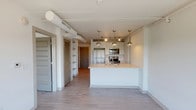 This 1-bedroom, 1-bathroom unit showcases a contemporary industrial-chic aesthetic, thoughtfully designed to maximize both function and style. The open-concept living area features light wood-style flooring that flows seamlessly throughout the space, complemented by crisp white walls and exposed ductwork along the ceiling that adds an urban, loft-inspired character.

The kitchen serves as the centerpiece of the main living area, anchored by a sleek peninsula with a stainless steel undermount sink, white cabinetry with brushed nickel hardware, and light quartz-style countertops. Pendant lighting illuminates the workspace, while a suite of stainless steel appliances — including a full-size refrigerator, range, microwave, and dishwasher — provides a complete culinary setup. A dedicated open shelving unit nearby offers additional storage and display space.

The bathroom is finished with clean, modern details, including white vanity cabinetry, a large mirror, quartz countertop, and a glass-enclosed walk-in shower. Light flooring ties the bathroom aesthetic cohesively to the rest of the unit.

Track lighting and pendant fixtures throughout the unit ensure well-distributed illumination, while windows bring in natural light and views of the surrounding area, enhancing the overall bright and airy ambiance of this efficiently designed home.