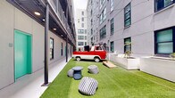 This urban outdoor courtyard is a thoughtfully designed communal amenity space tucked between the facades of surrounding multi-story residential buildings. The ground is covered in lush artificial turf that runs the length of the elongated corridor-style courtyard, creating a refreshing green focal point in an otherwise urban environment. String lights are draped overhead across the full span of the space, adding a warm, inviting ambiance during evening hours. A eye-catching vintage red-and-white truck serves as a bold centerpiece and conversation piece, fitted with what appears to be audio equipment, reinforcing the space's social character. Recreational amenities include a full-size ping-pong table and a cornhole board, encouraging casual, active use by residents. Seating is casual and eclectic, featuring striped pouf ottomans and small round side tables, as well as wire accent chairs arranged around a bistro table at one end. Large, modern white rectangular planters line one end of the courtyard, filled with ornamental grasses and tropical-style plants, adding natural texture and greenery. On one side, a covered walkway with recessed lighting provides a sheltered passage, accented by teal-painted doors that add a vibrant pop of color. The overall atmosphere is lively, modern, and community-focused.