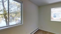 This two-story space is characterized by clean, minimalist interiors finished in crisp white walls and neutral-toned carpeting. The image highlights a well-proportioned room featuring a large, modern casement window that floods the space with natural light while framing a lush outdoor view of mature trees and what appears to be a tranquil waterway beyond. The window's generous size enhances the sense of openness and brings the surrounding landscape directly into the interior. A baseboard heating unit runs along the lower wall beneath the window, providing a practical and unobtrusive heating solution. The room's simple design and smooth wall surfaces contribute to a clean, functional aesthetic throughout. Spread across two floors, the overall layout offers versatility in how the space can be utilized, allowing for clear separation between living and other functional areas. The neutral palette and abundant natural light create a bright, airy ambiance, while the scenic outdoor views add a calming, natural element to the interior environment.