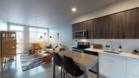 This single-floor residence showcases a cohesive contemporary design with polished concrete flooring, recessed lighting, and a neutral palette carried throughout the space. The open-concept kitchen and living area form the heart of the unit, where the kitchen impresses with dark wood upper cabinetry contrasted against white lower cabinets, a white quartz countertop, stainless steel appliances including a French door refrigerator, electric range, and built-in microwave. A concrete-topped island with bar seating provides a natural transition into the dining and living areas. A sliding barn door adds a rustic accent near the kitchen. The living area benefits from large windows that draw in generous natural light and features polished concrete floors that extend throughout. The bedroom offers carpeted flooring, soft wall tones, and direct access to a walk-in closet with built-in shelving and hanging rod. The bathroom is finished with a floating white vanity, large mirror, and polished concrete floors. A dedicated den serves well as a home office, complete with a window, closet, and concrete flooring, rounding out this well-appointed, thoughtfully designed space.