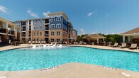 Located at 4964 NC-55 Hwy in Cary, North Carolina, this outdoor amenity space offers a resort-inspired experience designed for relaxation and community gathering. At its centerpiece is a generously sized swimming pool with a curving, freeform shape and crystal-clear water, surrounded by a broad concrete pool deck lined with lounge chairs for sunbathing. Accessibility features, including a pool lift, ensure the space is welcoming to all.

Adjacent to the pool is a covered pavilion constructed with striking stacked stone columns and warm wood-beamed ceilings, creating an inviting indoor-outdoor atmosphere. The pavilion is outfitted with a full outdoor kitchen setup, complete with built-in appliances set into the stone wall, as well as a wall-mounted television — ideal for entertaining. Comfortable seating, including hanging egg chairs, dining tables, and benches, fills the shaded space, while ceiling fans provide a cool breeze on warm days.

The pavilion opens directly toward the pool, offering unobstructed views of the water and the lush tree line beyond. The multi-story residential building, clad in a combination of blue siding and warm brick, serves as a backdrop, enhancing the cohesive architectural character of the complex. The overall ambiance is polished, social, and resort-like, making this outdoor space a premier communal destination.