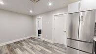This efficiently designed single-floor unit offers a thoughtful layout that maximizes every square foot. The open kitchen and living area serves as the heart of the space, featuring stainless steel appliances including a French-door refrigerator, electric range, microwave, and dishwasher. White shaker-style upper cabinets contrast elegantly with dark lower cabinetry, while a geometric hexagonal tile backsplash adds a modern, decorative touch. Light gray wood-look flooring runs throughout the unit, creating a cohesive and contemporary aesthetic. The bedroom is a bright, well-proportioned room with a sliding window that brings in natural light and offers a view of the surrounding exterior. Neutral gray walls and clean white trim maintain the unit's modern feel. The bathroom is finished to a high standard, showcasing large-format marble-look floor tiles, a subway-tiled walk-in shower with a glass sliding door and built-in niche, and a dark-toned floating vanity. A dedicated laundry area adds everyday convenience. Recessed lighting throughout the unit ensures consistent, clean illumination. Overall, the space presents a polished, move-in-ready interior with modern fixtures and a neutral palette.