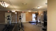 This single-floor fitness room offers a practical and functional workout environment designed to support a range of exercise needs. The space is finished with warm, earth-toned walls that create an inviting atmosphere, complemented by striped carpeting underfoot that adds comfort during workouts. Ceiling-mounted light fixtures provide consistent overhead illumination throughout the room, while a window with blinds allows for natural light to filter in.

The room is equipped with key cardio machines, including an elliptical trainer and a treadmill positioned on a protective floor mat. A wall-mounted equipment rack provides additional options for strength training, and free weights are also accessible within the space. Full-length mirrors along one wall enhance the sense of openness and allow users to monitor their form during exercise.

Ventilation is provided through ceiling ducts, ensuring airflow throughout the room. A clearly marked emergency exit door is visible, reflecting attention to safety standards. The overall layout is efficient and well-organized, making good use of the available footprint to accommodate multiple users and workout styles simultaneously. This fitness room serves as a convenient, well-appointed space for maintaining an active lifestyle.