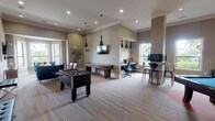 This single-floor Social Room is thoughtfully designed to serve as a versatile gathering space that balances leisure and functionality. The open layout is anchored by warm, neutral-toned walls, crown molding, and a combination of hardwood-style flooring and patterned carpeting that defines each activity zone. A lounge area near the entrance features a vibrant teal velvet sofa and coordinating seating arranged around a geometric coffee table, with a wall-mounted flat-screen TV overhead. Sleek black pendant lighting fixtures are suspended throughout, adding a modern, sophisticated touch to the space. The game area is a standout highlight, equipped with a foosball table, a shuffleboard table, and a dedicated billiards room. The billiards room features a blue felt pool table centered beneath a contemporary globe chandelier, with a distinctive decorative wall display of varied clocks adding artistic character. A built-in workspace nook accommodates multiple workstations with ergonomic seating, desktop monitors, and tall bar-height seating along a counter, all illuminated by natural light streaming through large windows with outdoor views of lush greenery. Recessed lighting throughout ensures the entire space remains bright and welcoming at all hours.