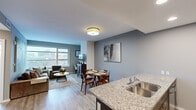This well-appointed 3-bedroom, 1-bathroom unit offers a thoughtfully designed open-concept layout across a single floor. The kitchen is outfitted with dark espresso cabinetry, granite countertops, a double stainless steel sink, a tile backsplash, and a full suite of stainless steel appliances including a refrigerator, range, and microwave. A granite-topped island seamlessly connects the kitchen to the dining area, where wood-toned flooring flows throughout the main living spaces. The dining area accommodates a full table and chairs beneath neutral gray accent walls. The adjacent living room is bright and airy, featuring large floor-to-ceiling windows that frame expansive urban skyline views, allowing abundant natural light to fill the space. The primary bedroom continues the city view theme with a wide window overlooking the surrounding cityscape and includes closet space and plush carpeting. The shared hallway connects the bedrooms to the rest of the unit with clean, modern finishes including recessed lighting and white paneled doors. The full bathroom features a granite-topped dark wood vanity, tile flooring, and a shower-tub combination. Overall, the unit presents a contemporary urban aesthetic with a clean, neutral palette throughout.
