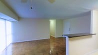 This single-floor space presents an open, functional layout with a clean and straightforward design. The living area is characterized by classic parquet wood flooring arranged in a geometric block pattern, adding warmth and texture to the room. The walls are finished in a neutral off-white tone, creating a bright and versatile backdrop that complements a variety of interior styles. The ceiling is smooth and painted in a soft, cool tone, with a ceiling fan fixture providing practical overhead airflow.

A partial kitchen peninsula with a dark granite-style countertop edges into the living space, offering a natural division between the kitchen and main living area while maintaining an open feel. This counter feature provides functional prep or casual dining space.

A hallway extends from the main living area toward the front entrance, where a white door with a standard handle is visible. The hallway is lit by a wall-mounted light, ensuring the transition space remains well-illuminated. The overall ambiance of the space is simple and uncluttered, offering a neutral foundation that can be personalized with furnishings and décor.
