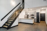 Unit 110 at 11800 Main St in Fredericksburg, Virginia is a two-story live-work space that masterfully combines industrial aesthetics with contemporary design. The main level features polished concrete floors, exposed brick accent walls, and large black-framed windows that invite abundant natural light into the open living area. The kitchen is a standout, showcasing a central island with a granite countertop, an integrated sink with a modern faucet, pendant lighting overhead, and a full suite of stainless steel appliances including a refrigerator, range, microwave, and dishwasher — all complemented by gray shaker-style cabinetry. A sleek staircase with a bold dark frame and glass railing connects the two floors, adding a striking architectural focal point. The lower level also features a full bathroom with a tiled tub-and-shower surround, granite-topped vanity with an undermount sink, and clean, neutral tile flooring. The upper level offers hardwood-style flooring, crisp white walls, and a pass-through interior window that overlooks the main floor below, enhancing the open, connected feel of the space. The upper level also includes a well-appointed bathroom with a granite vanity. Throughout, bold black trim and door frames create a cohesive, modern industrial look that defines the character of this distinctive two-story unit.